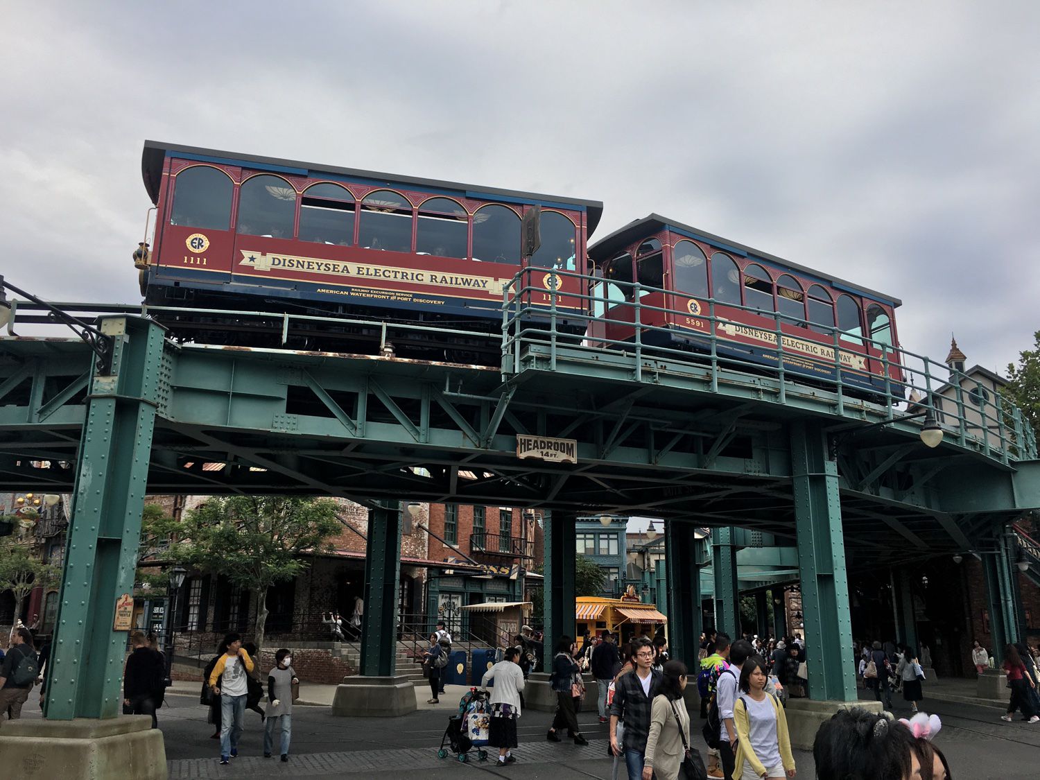 A train set on the Tokyo DisneySea Electric Railway passing over the elevated tracks in American Waterfront while people walk beneath it.